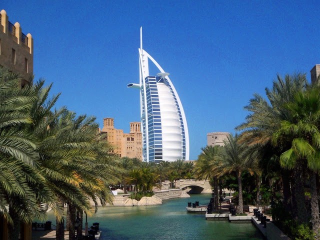 The Burj al Arab hotel, a white building shaped like a nillowing sail, surrounded by palm trees and water