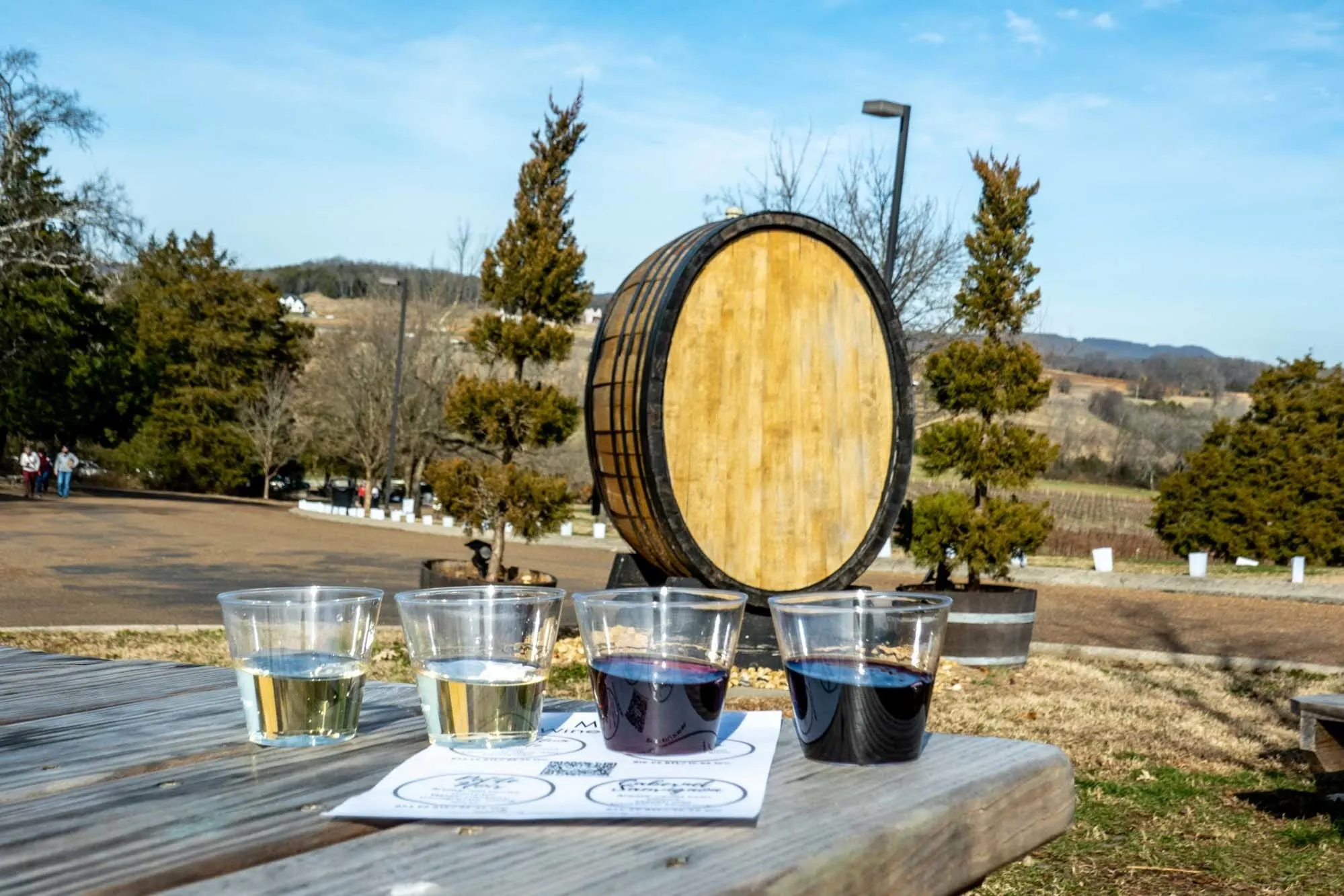 Wine tasting flight on a picnic table beside a large wooden barrel.